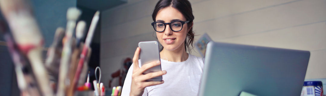 Image of young woman sitting at a laptop computer, holding and looking at a cell phone