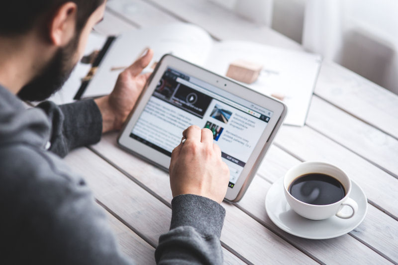 Image of man holding and looking down at electronic tablet, with a cup of coffee to its right