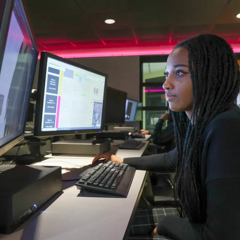 Photo of a young woman looking at two computer screens with red lighting in the background