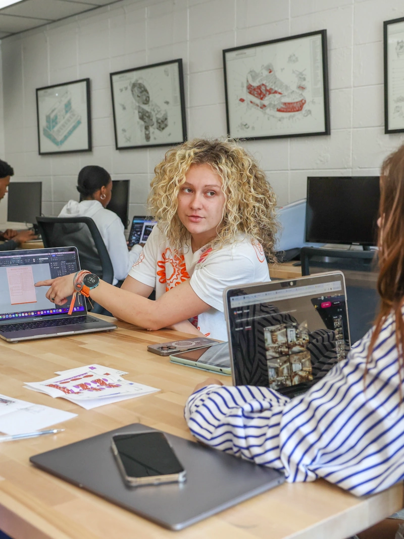 A young woman sits at a table, pointing to something on her computer screen, showing it to another young woman who is partially off-camera in the foreground