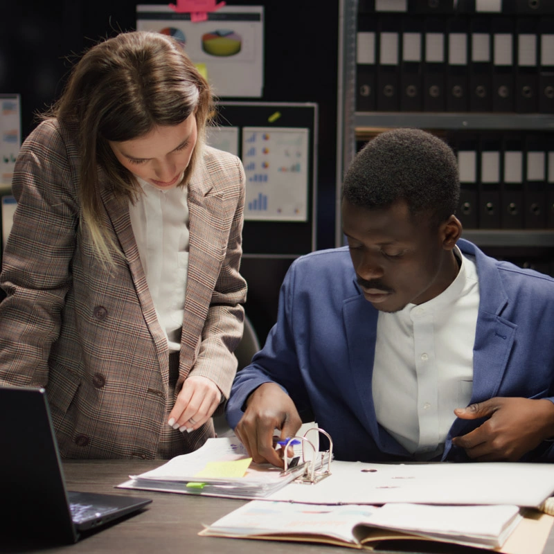 A standing man and sitting woman look at a two-ring binder