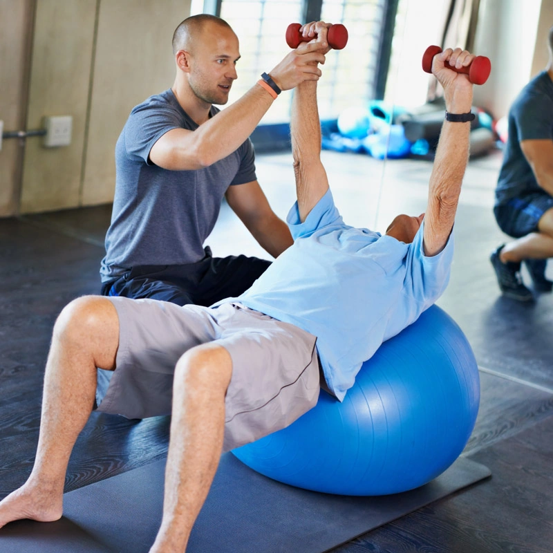 A physical therapist helps someone exercise with hand weights and an exercise ball