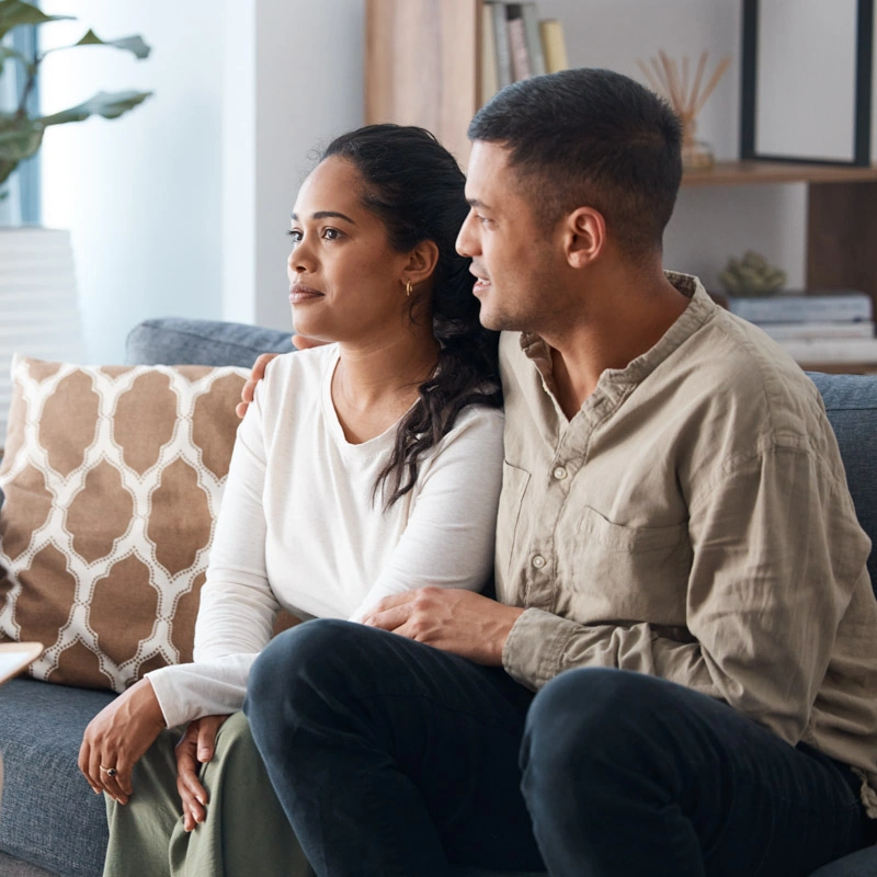 A young man and woman sit on a couch; the young man has his arm around the young woman's shoulder