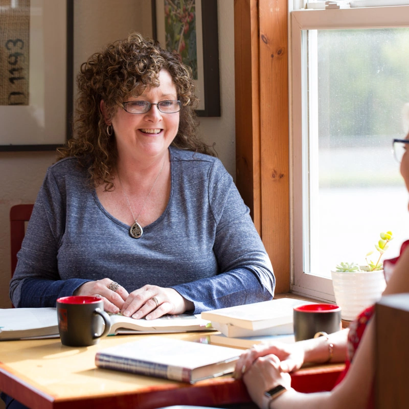 A woman sits at a desk and smiles at an out-of-focus woman in the foreground on the other side of the desk