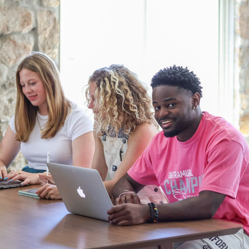 Three students sit at a table as the closest, an African-American man in a bright pink shirt, looks at the camera and smiles