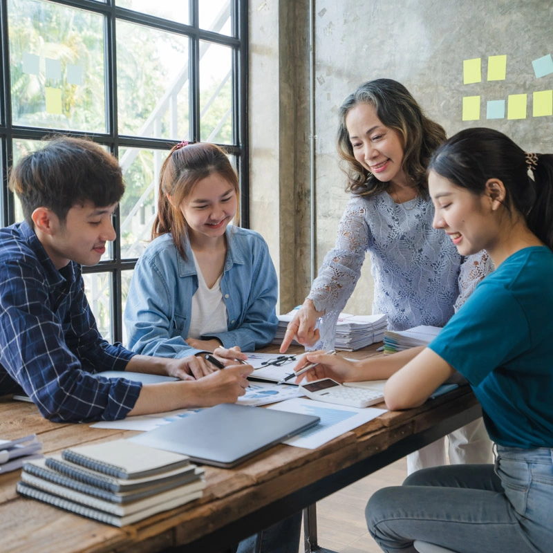 An adult woman gestures at a textbook on a table as three college-aged students look at the textbook