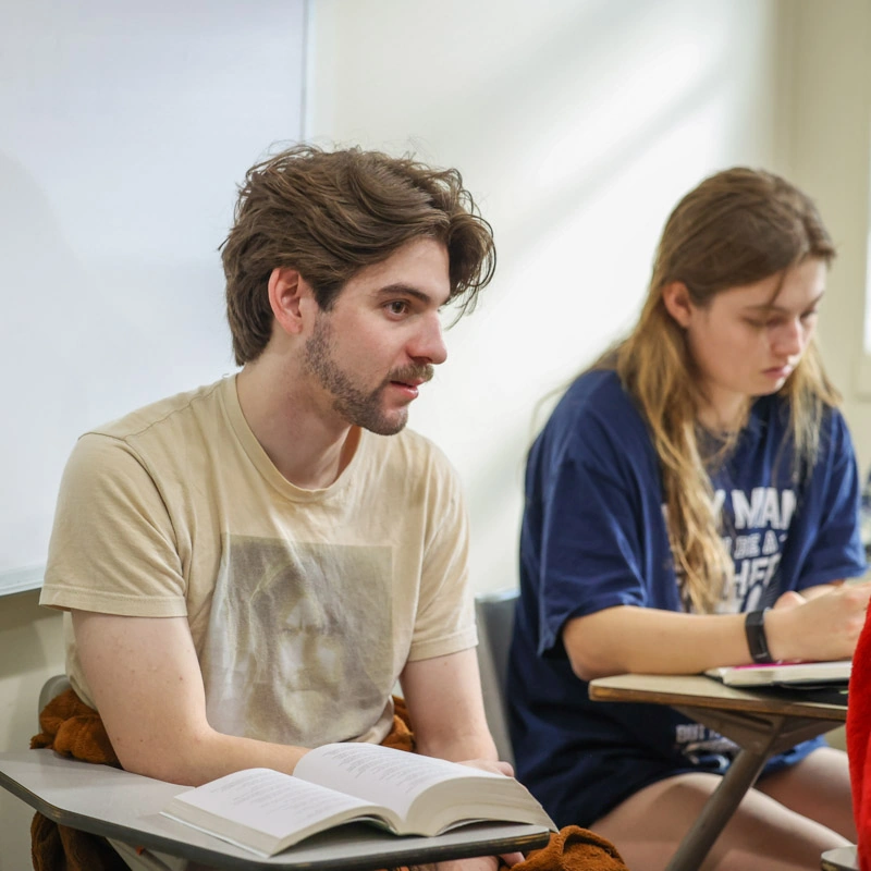 A young man sits at a college desk; the desk holds a very large textbook