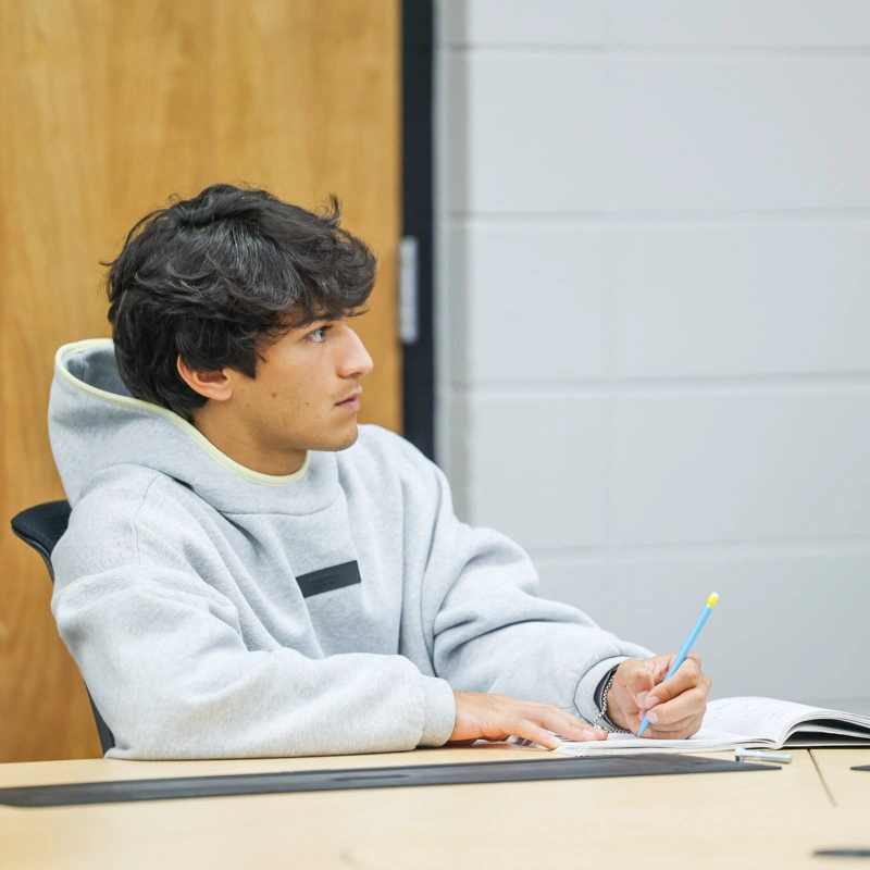 A young man sits at a college desk, writing in a notebook