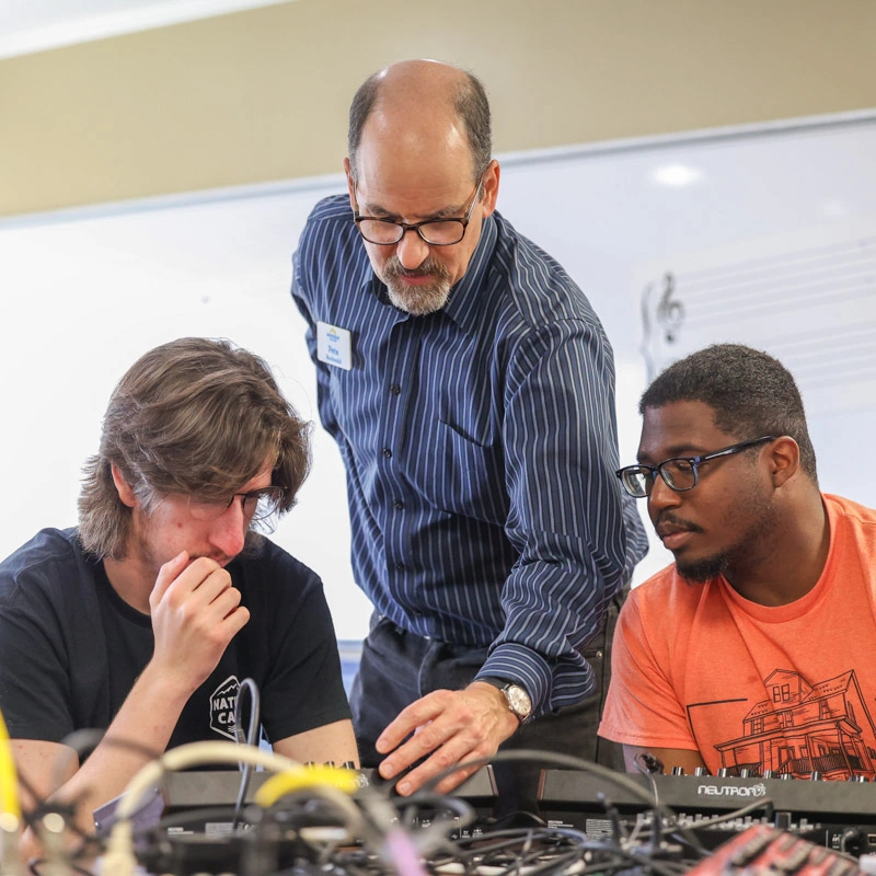 A standing male music business professor shows two seated male college students how to adjust the dials on a mixer