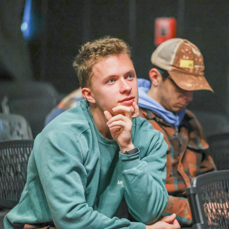 A young man leans forward, looking pensive, holding his chin in his hand