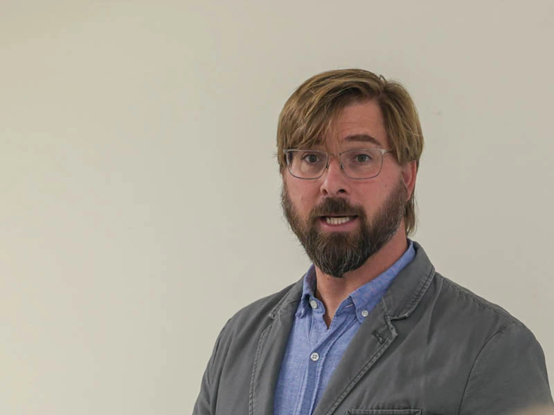 A middle-aged man talks in front of a white wall