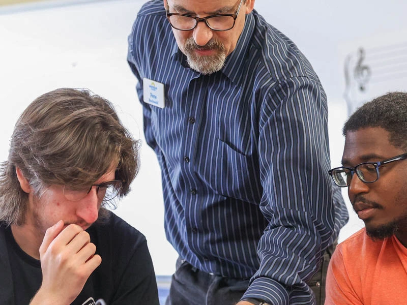 A standing college professor leans over and points something out to two seated college students