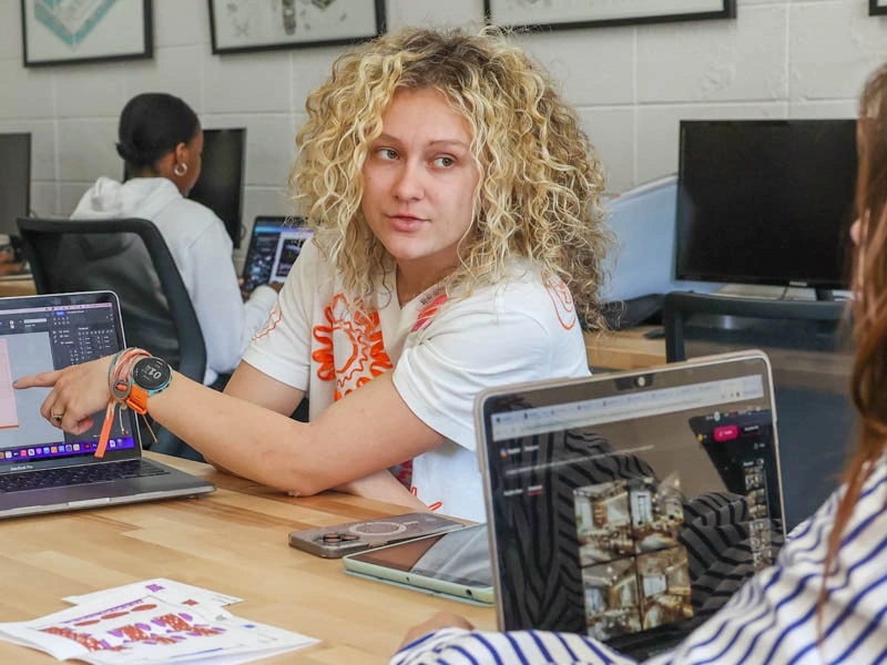 A young woman points to her computer screen while talking to someone else