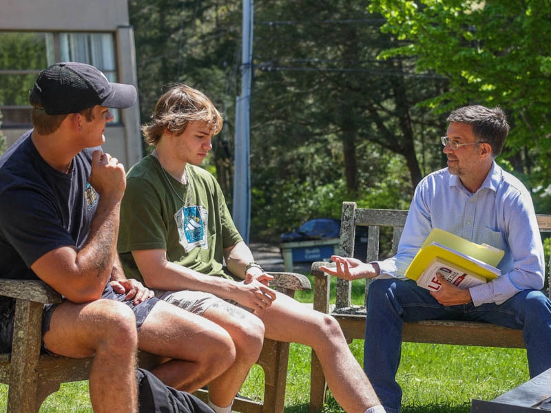 A professor talks with two young men as they sit outside on benches