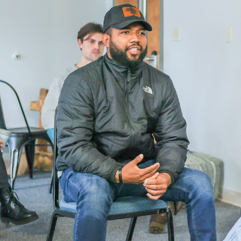 A young man sits in a stackable chair while wearing a jacket and jeans