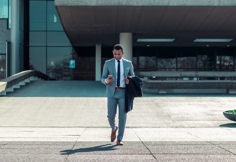 A young man in a suit walks outside, looking down at his cell phone