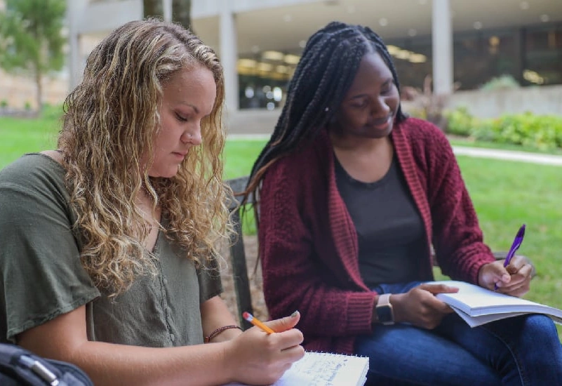 Two young women write on notebooks while sitting on a bench outside the L. Nelson Bell library