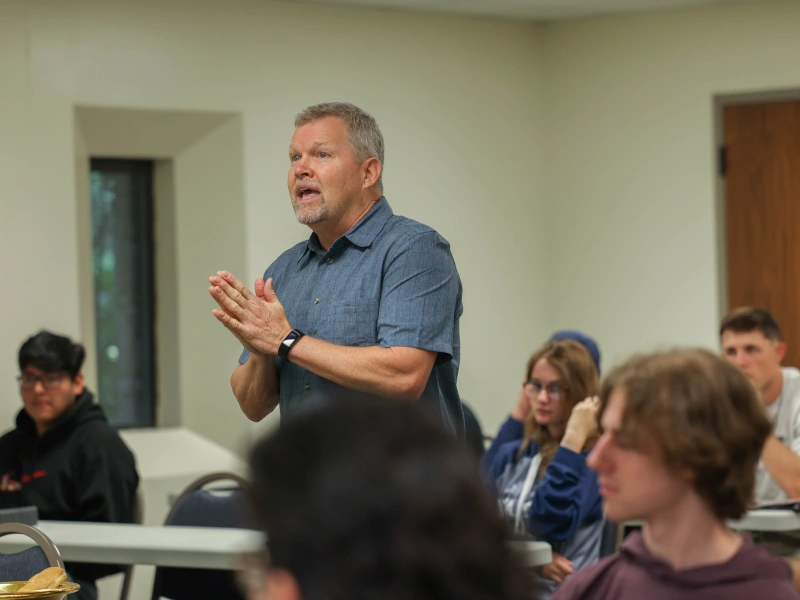 A man with hands pressed together stands and speaks in a classroom