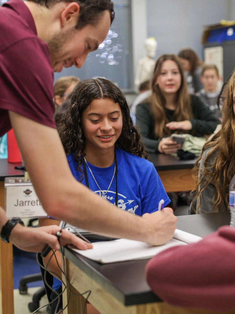 A young man in a classroom assists a girl who is seated at a desk