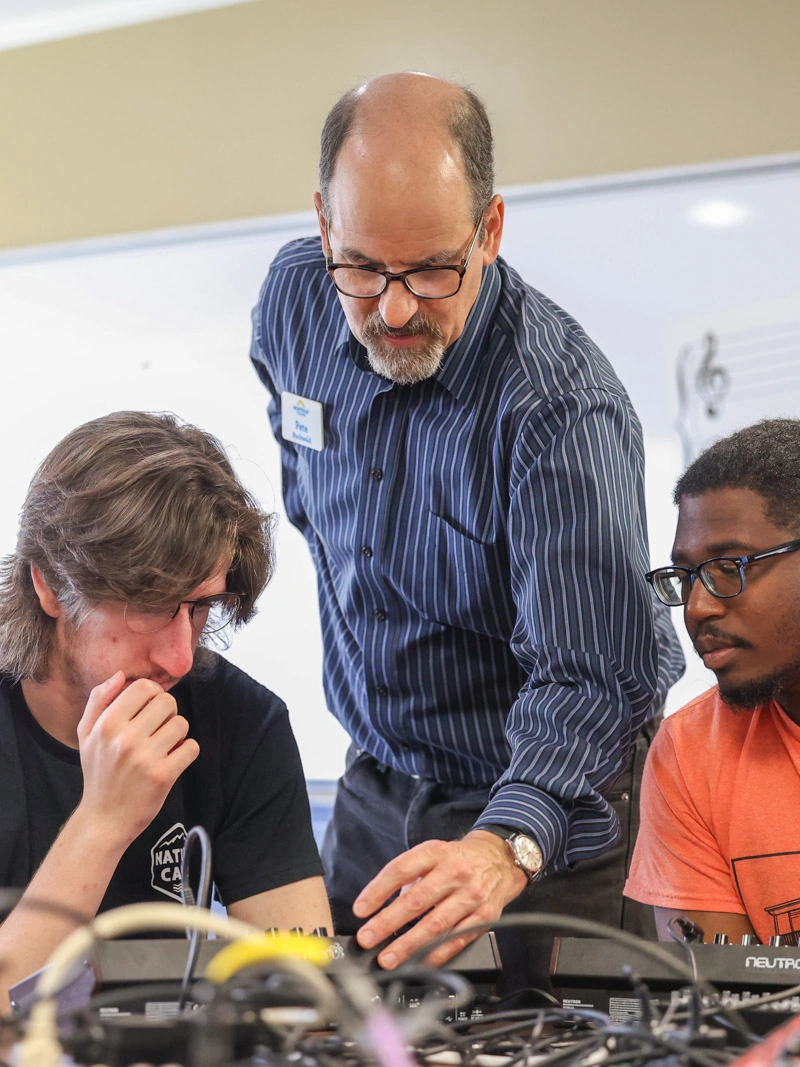 A male professor shows two male students how to change settings on an audio mixer