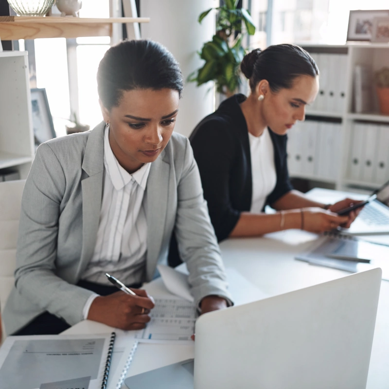 Two women in business suits sitting at a desk working on paperwork